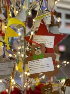 a close up photo of one of the giving trees focusing on a brown paper ornament in the shape of a bear, with white Christmas lights lit and other paper ornaments around
